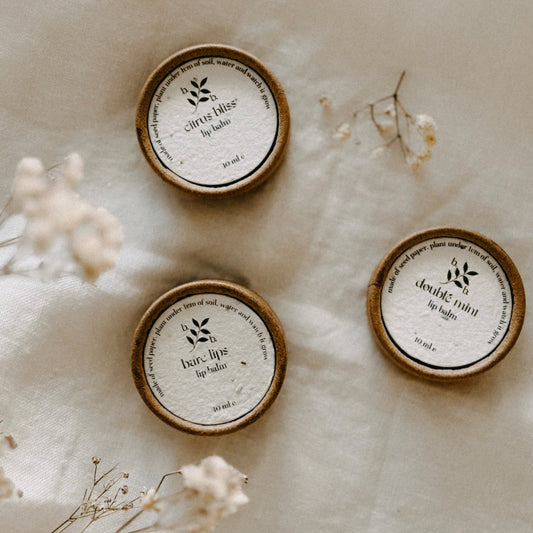 Three Bare & Bloom natural lip balms—Citrus Bliss, Bare Lips, and Double Mint—are displayed on a soft, neutral fabric, accompanied by delicate dried flowers. The biodegradable packaging and seeded labels reflect the brand's commitment to sustainability and natural skincare.