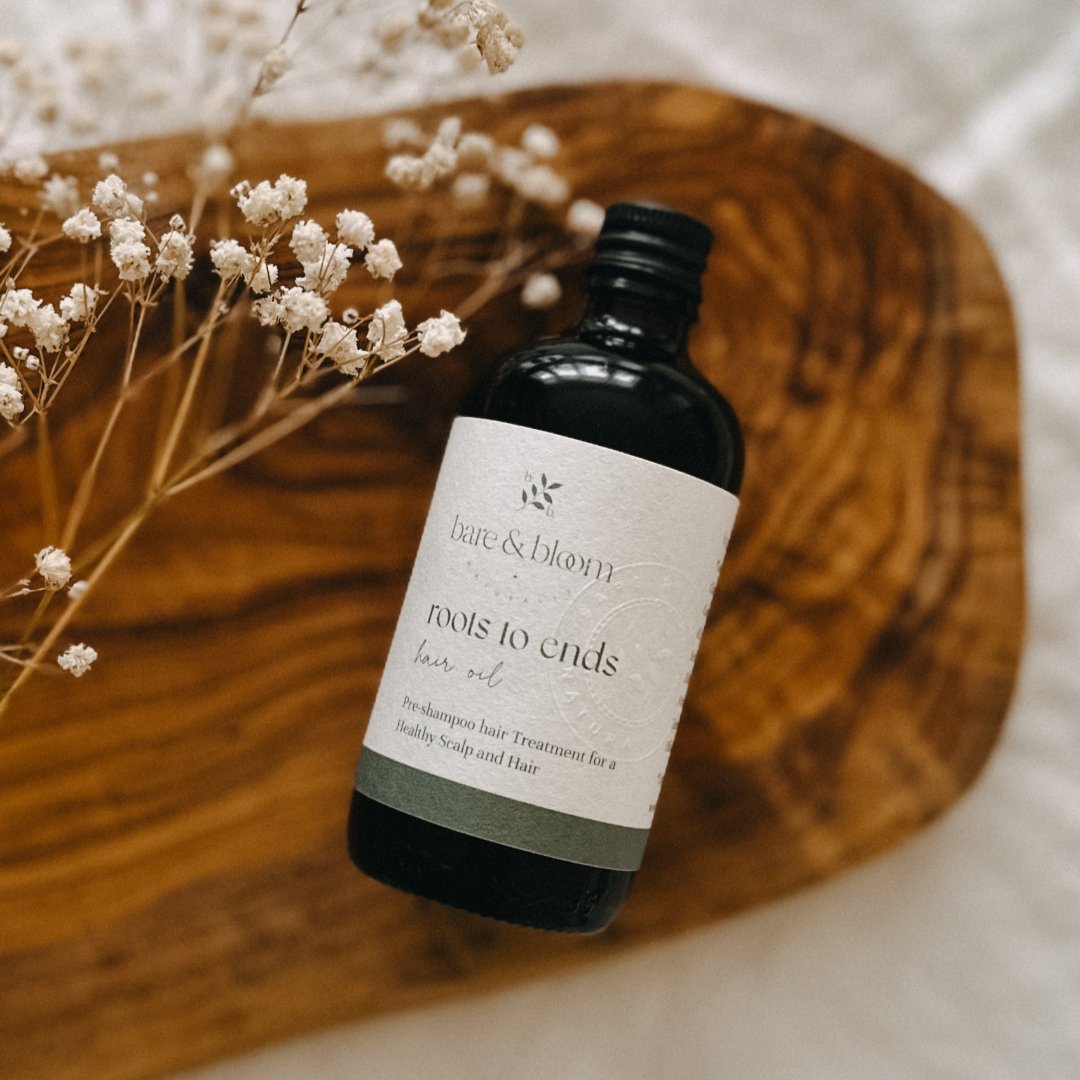 A bottle of 'Bare & Bloom Roots to Ends Hair Oil' rests on a wooden surface, surrounded by delicate dried flowers. The bottle is labeled as a pre-shampoo hair treatment for a healthy scalp and hair.