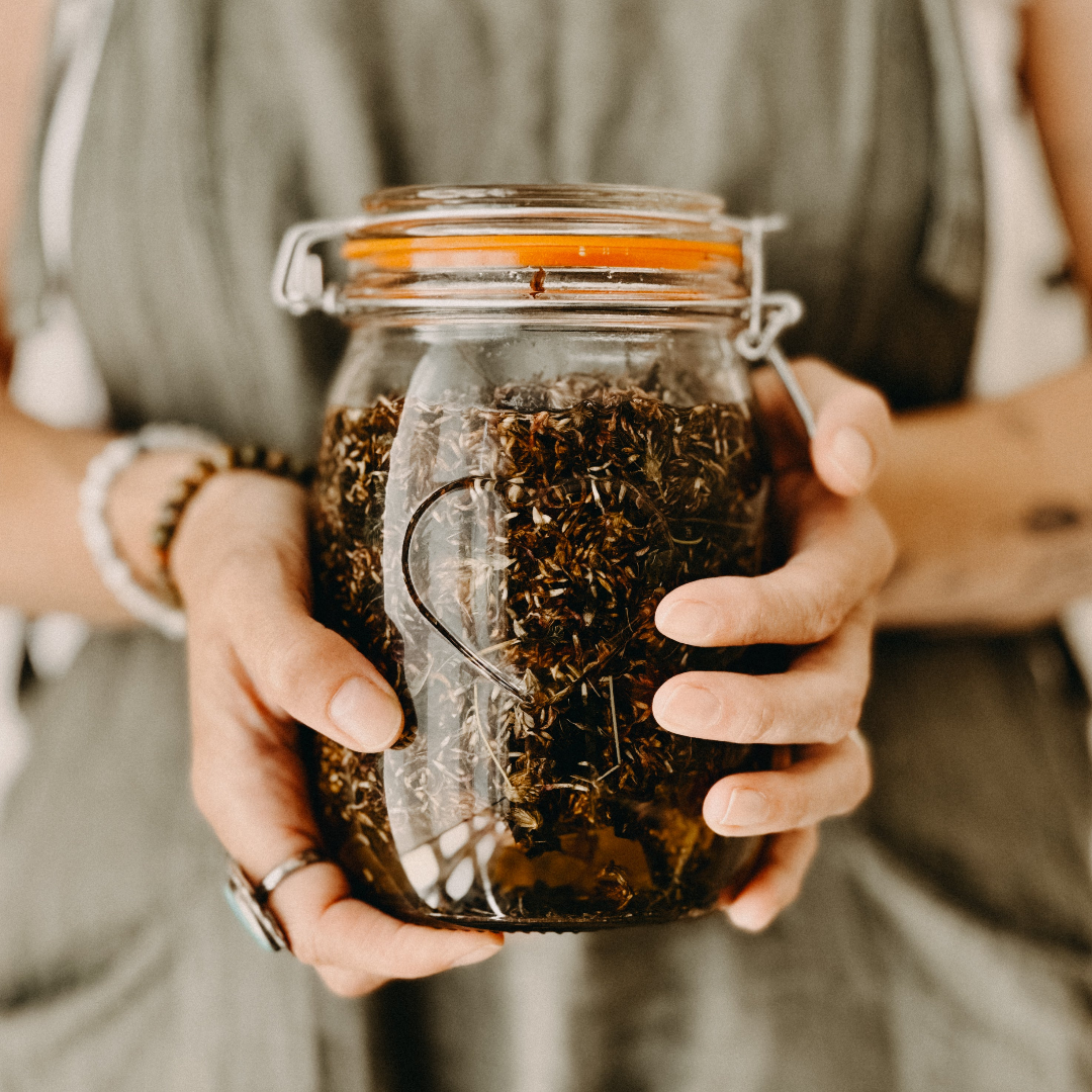 Hands holding a glass jar filled with dried herbs or tea leaves. The jar has an orange clip-top lid.