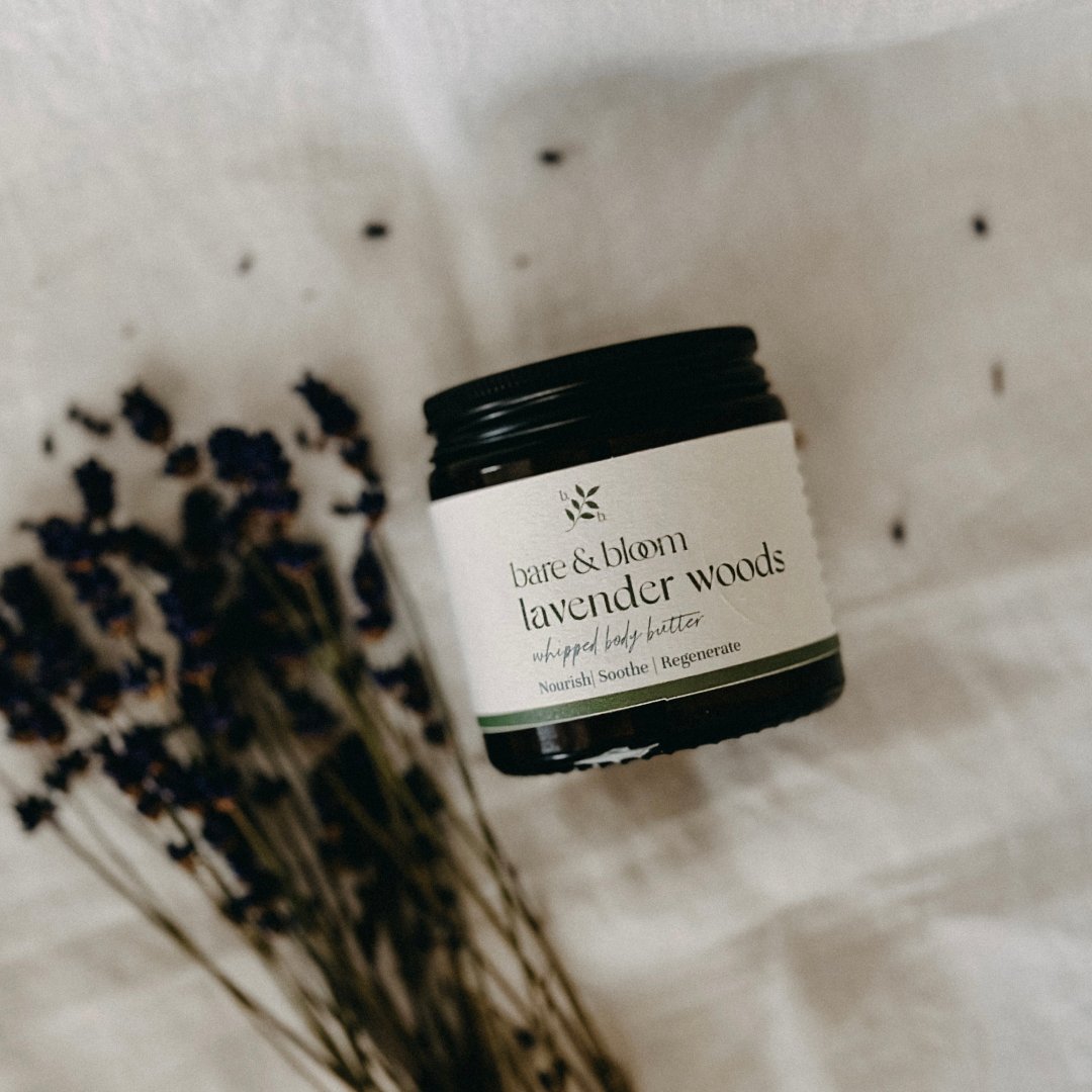 A jar of Bare & Bloom Lavender Woods Whipped Body Butter is placed on a soft, natural fabric surface, surrounded by dried lavender sprigs. The jar features a minimalist label that highlights its nourishing, soothing, and regenerating properties, evoking a sense of calm and natural luxury